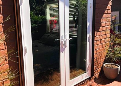 The newly completed door installation features white patio doors with glass panes that reflect greenery, set in a red brick wall on a wooden deck, accompanied by a potted plant.