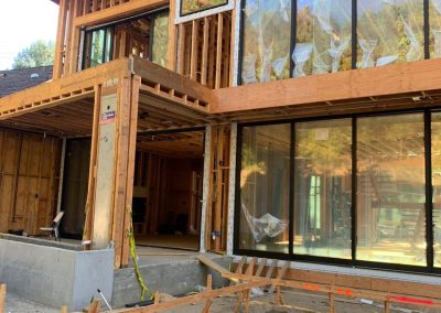 A partially constructed modern house with large glass windows, wooden framework, and a door installation that complements its sleek design.