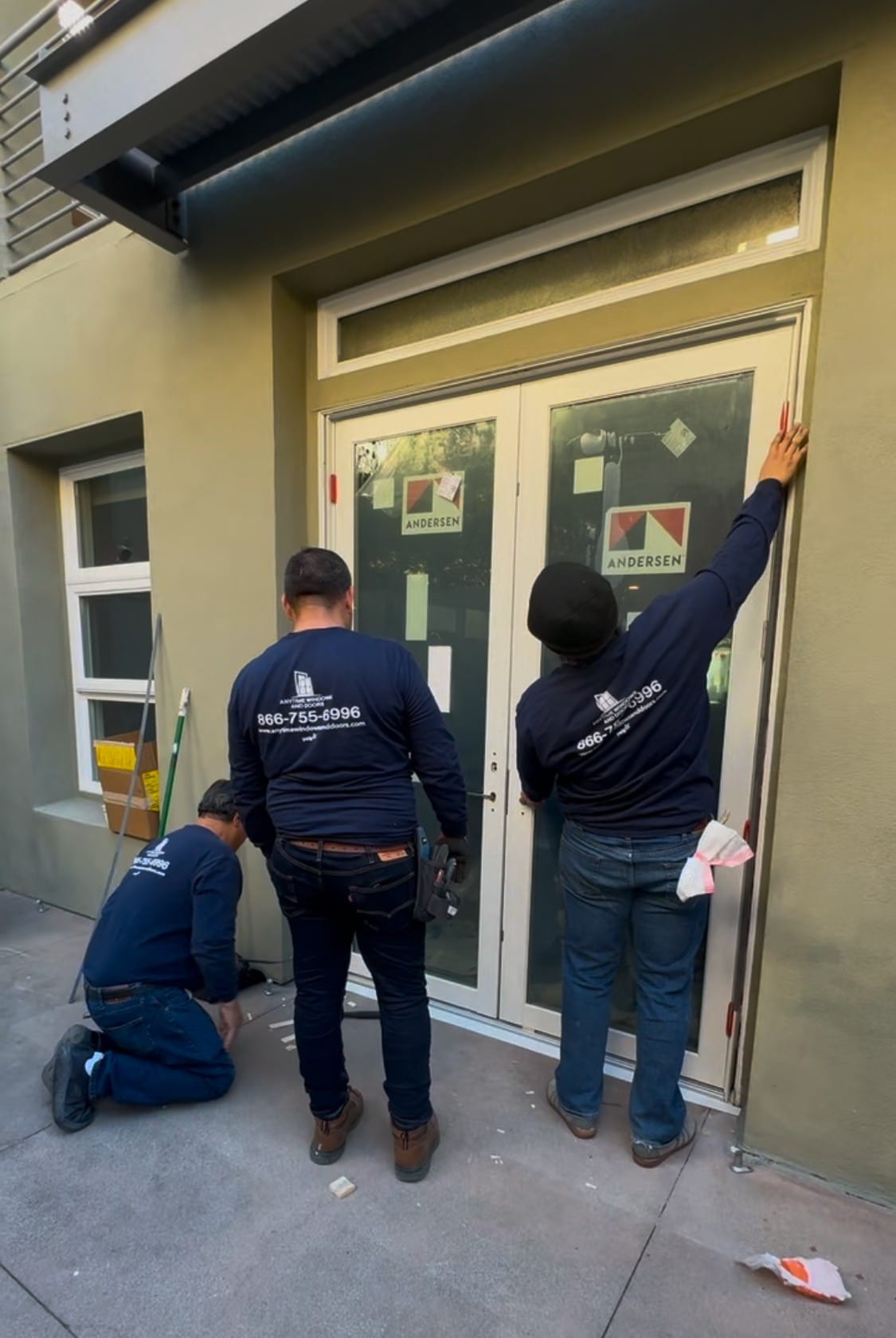 Item 1 Three workers in blue shirts install or inspect glass doors labeled "Andersen" on a building exterior.