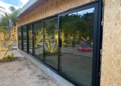 Large glass sliding doors reflect palm trees with string lights and a pink pool float outside a building under construction.