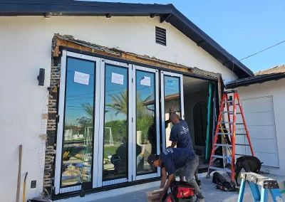 Two workers install large glass folding doors on a house; tools and a red ladder are nearby.
