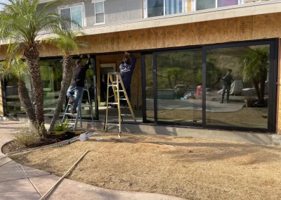 Two people on ladders install a window frame on a house exterior, with palm trees and dry grass nearby.