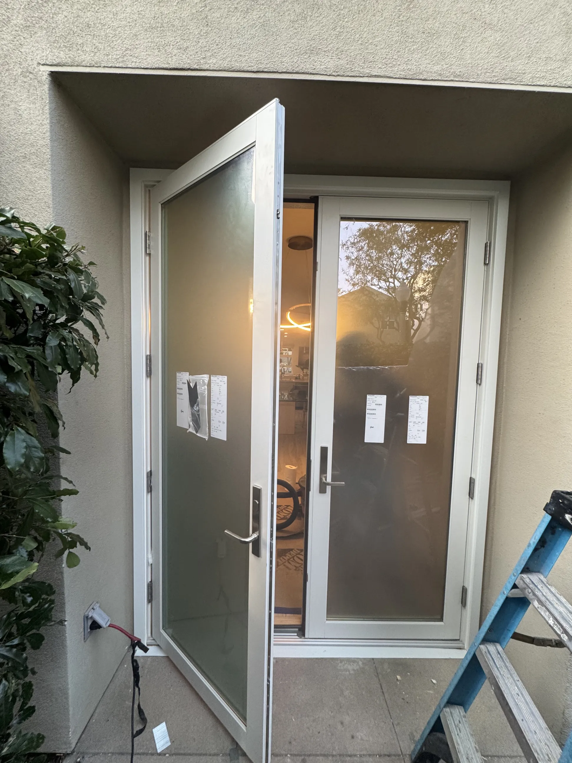 A glass patio door is partly open, with a ladder and some plants nearby, leading into a modern home.