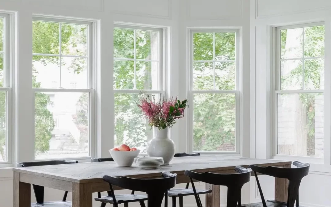 A bright dining room with large windows, a wooden table, black chairs, and a vase of flowers.